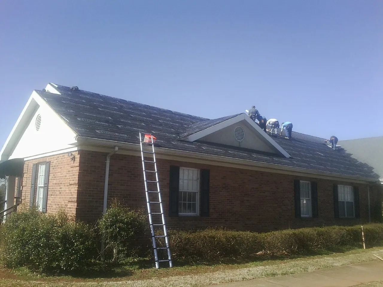 Crew tearing shingles off a brick ranch roof with a ladder set — Metal Roof Repair work by our crew in Uehling