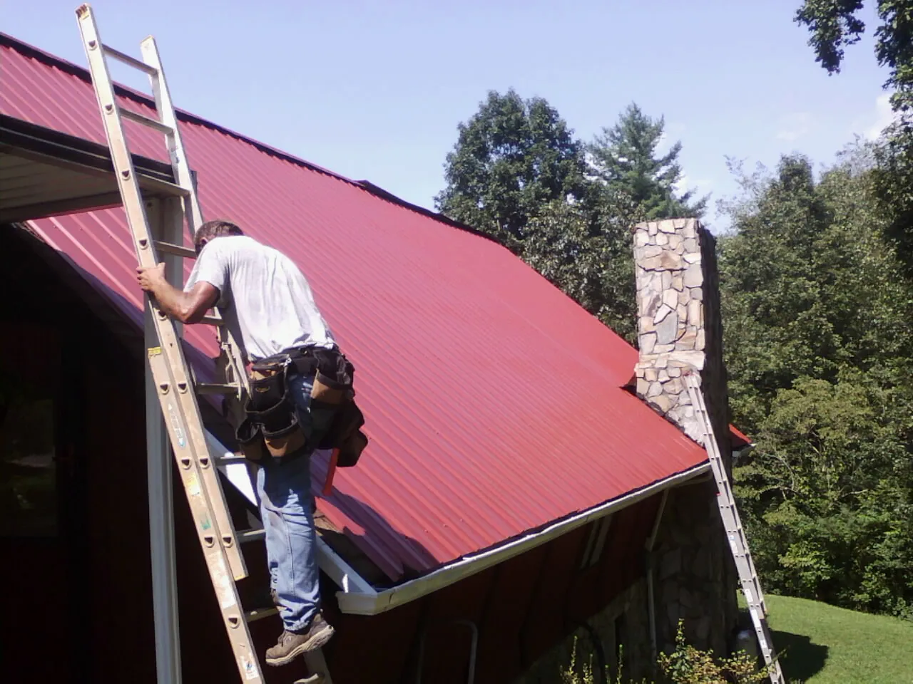Roofer on a ladder beside a red metal roof and stone chimney — Soffit & Fascia Repair by Roofing Brothers in Uehling